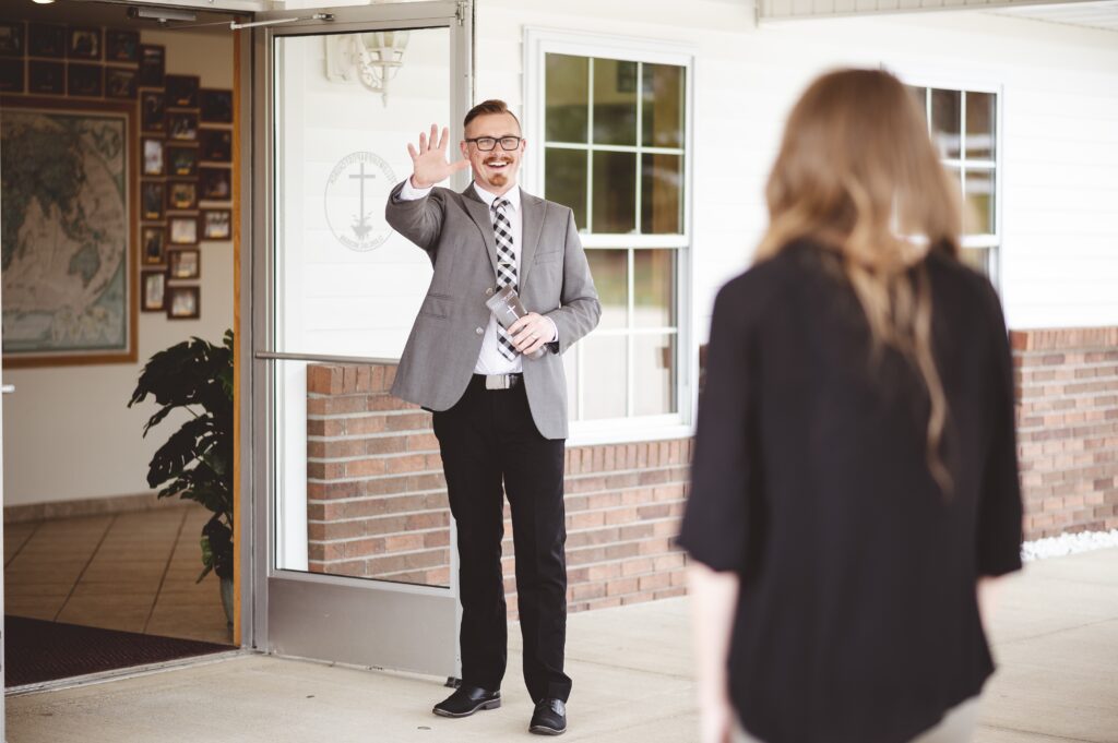 Pastor outside a church waving and welcoming a woman